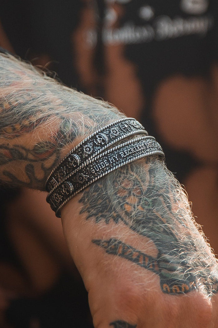 A tattooed forearm displays three handcrafted, dark metal bracelets with an adjustable fit. One reads GUIDE MY RECKLESS SOUL from Relentless Betrayal. A blurred figure in a black shirt enhances the mystique of the stainless steel design.