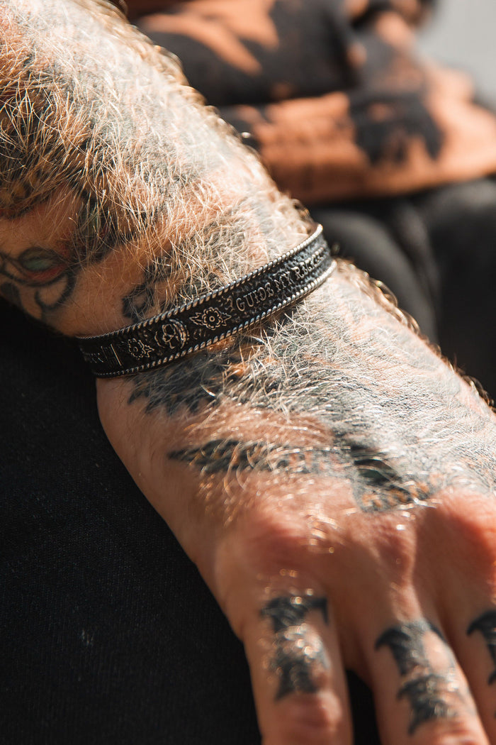 A tattooed forearm and hand display the Guide My Reckless Soul bracelet from Relentless Betrayal, featuring a handcrafted, textured design with engraved text. The adjustable stainless steel contrasts with intricate tattoos, while dark clothing completes the edgy look.