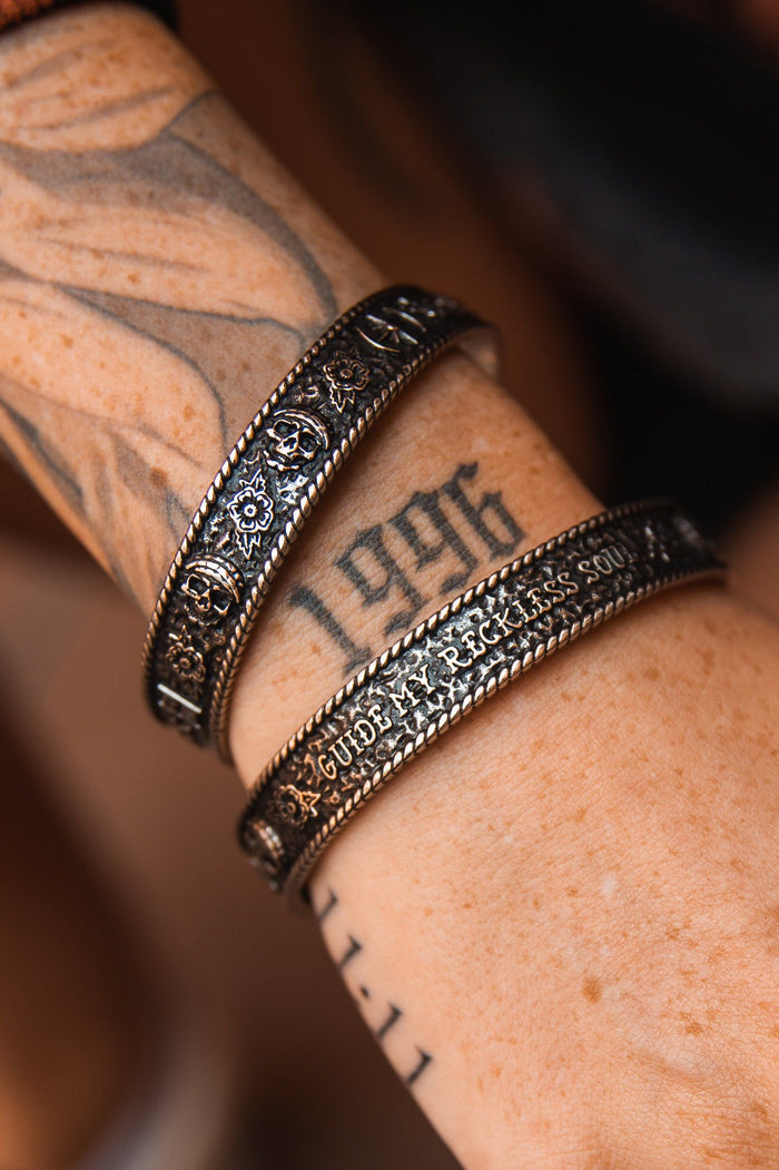 A close-up of a tattooed forearm with 1996 and GUIDE, adorned with two ornate, handcrafted metal bracelets. The Guide My Reckless Soul Bracelet by Relentless Betrayal features intricate patterns and textures, offering an adjustable fit for comfort.