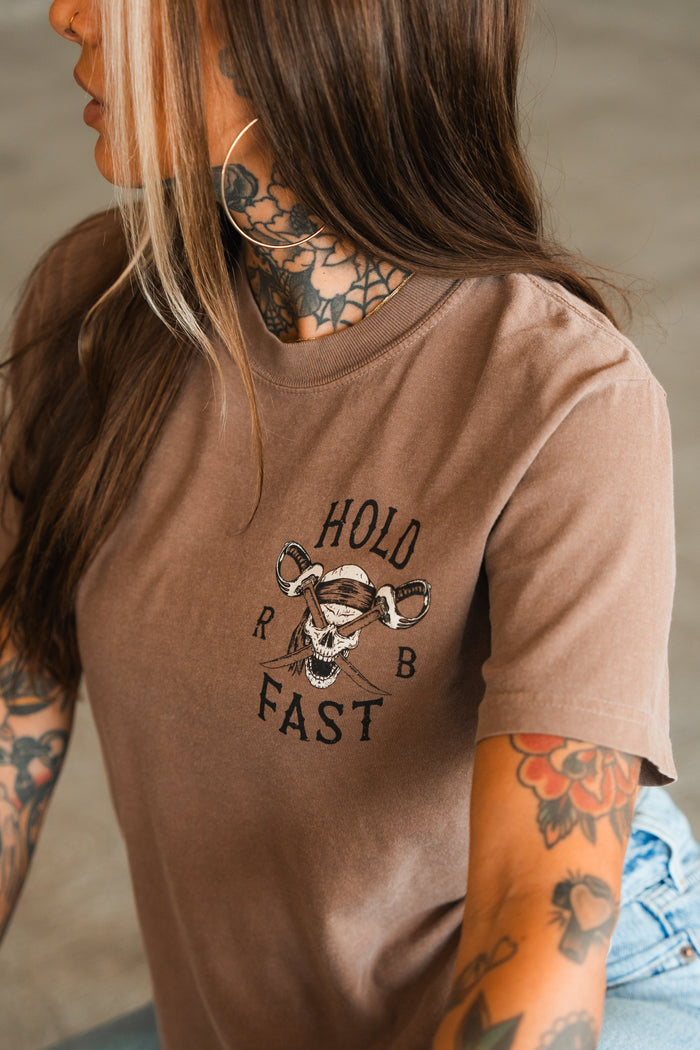 A person with long brown hair and tattoos wears the Relentless Betrayal Hold Fast Premium Cocoa T-Shirt, a heavyweight jersey cotton tee featuring a skull and crossed wrenches graphic with HOLD FAST printed on the front.