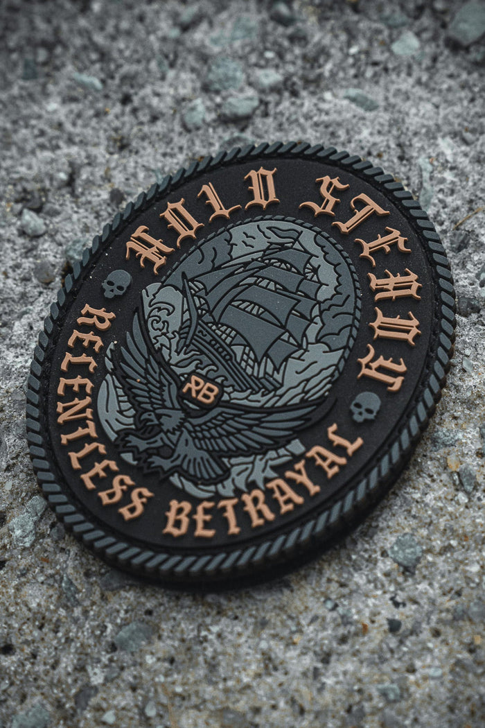 The Hold Steady Ghost Patch by Relentless Betrayal is a 3-inch round embroidered patch with a sailing ship, eagle, skulls, and "HOLD STEADY RELENTLESS BETRAYAL" in gold on black, shown on rough concrete.