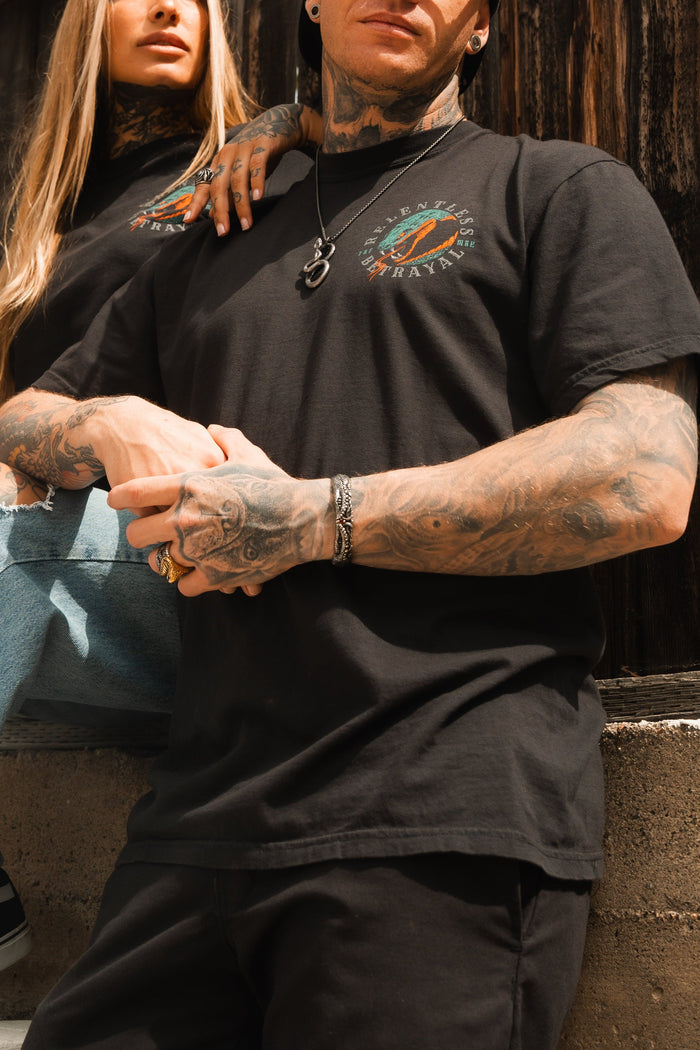 A tattooed man in a black Relentless Betrayal T-shirt with colorful graphics and a silver necklace stands by a wooden wall. Beside him, a tattooed woman, also in the matching T-shirt, rests her arm on his shoulder, displaying the stainless steel Holy Grail Bracelet by Relentless Betrayal.