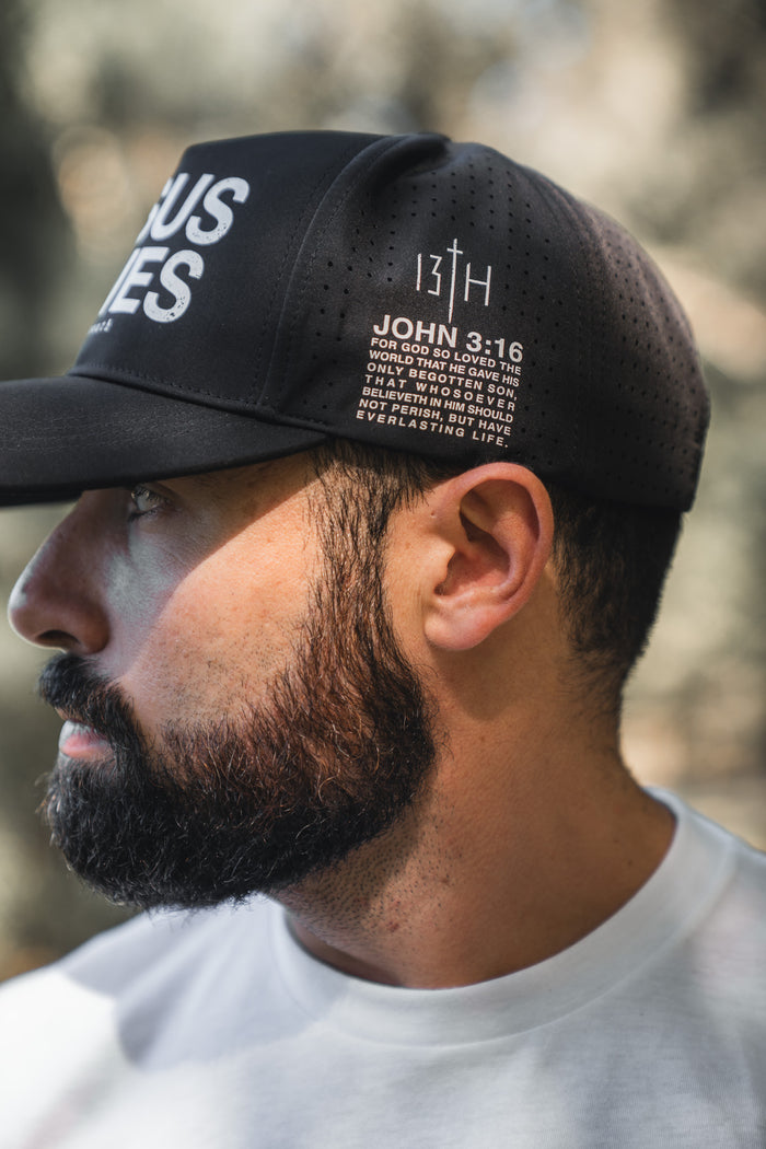 A bearded man in a white T-shirt wears the 13th Jesus Saves Black Snapback, featuring white perforated “JOHN 3:16” text, as he looks left outdoors with a blurred background.