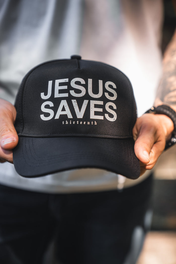 A person holds the Jesus Saves Black Snapback by 13th, featuring bold white "JESUS SAVES" text and smaller "thirteeneeth" lettering. They're dressed in a light shirt and dark pants.