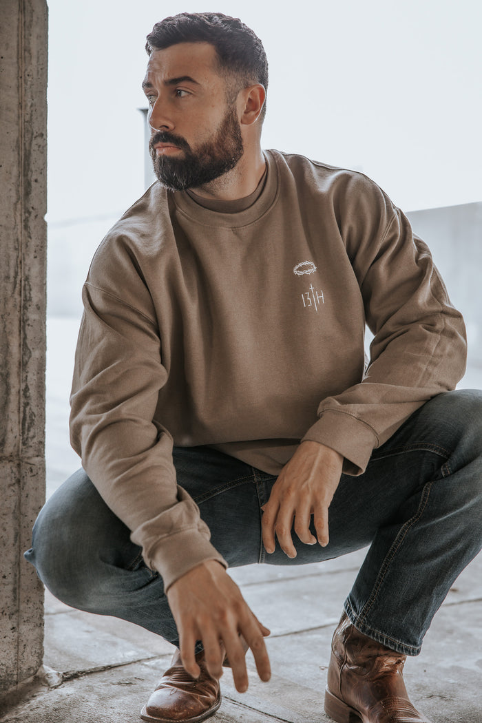 A bearded man squats near a concrete wall, looking left. He wears the Jesus Saves Crewneck by 13th, paired with blue jeans and brown boots. The bright background is slightly blurred.