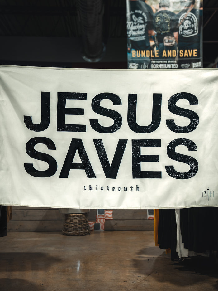 The Jesus Saves Flag - Ivory by 13th hangs indoors, featuring bold black text and “13TH” in the corner. Clothing racks and another sign appear in the background, enhancing the space with faith-inspired home decor.