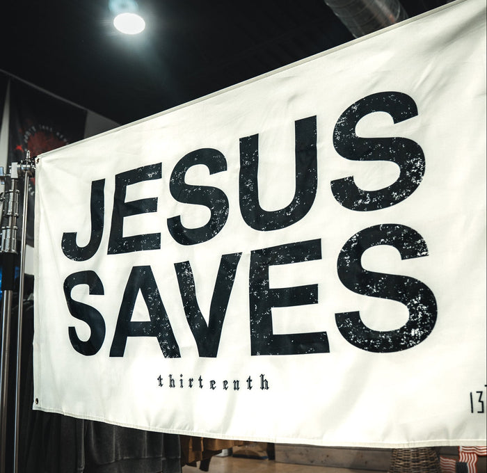 The Jesus Saves Flag - Ivory by 13th features bold, distressed black letters and "thirteenth" in small text below. Display it on a metal stand indoors for faith-inspired home decor or outdoors as a unique Christian statement flag.