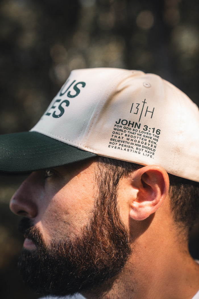 Close-up of a man wearing the 13th Jesus Saves Ivory Snapback, featuring “JOHN 3:16” and a Bible verse above his ear. The softly blurred background is lit with natural light.