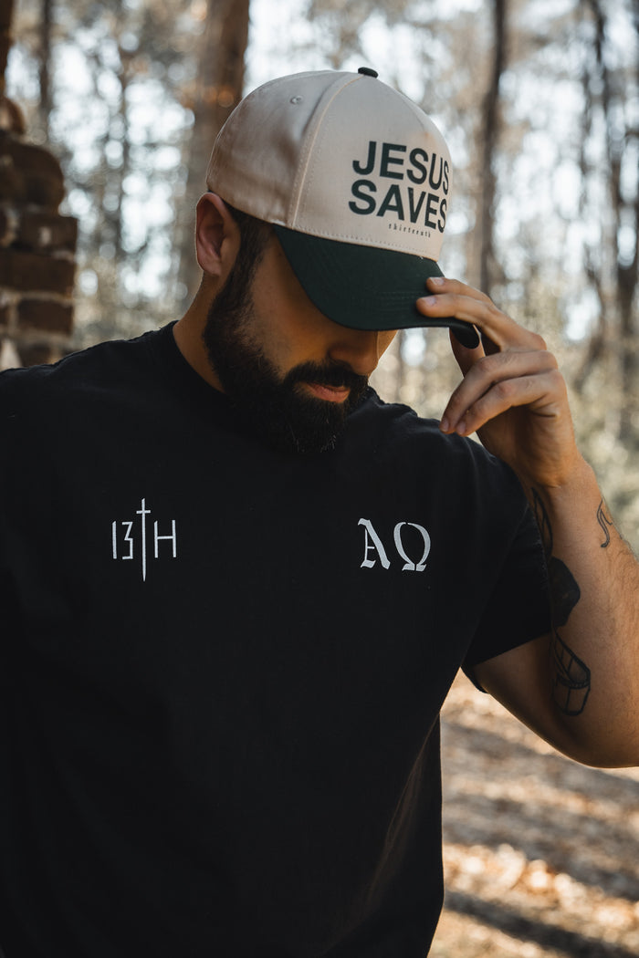 A bearded man stands outdoors by brickwork, wearing a black 13th t-shirt with Christian symbols and a Jesus Saves Ivory Snapback by 13th, holding its brim and looking down.