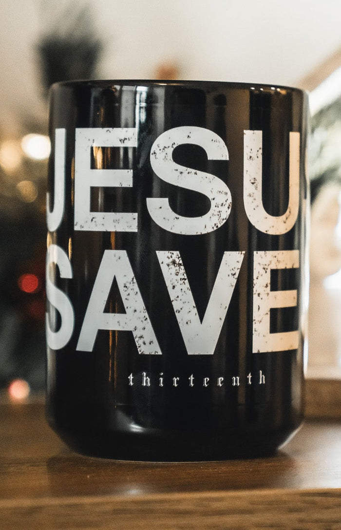 A black 13th Jesus Saves Mug with bold white lettering sits on a wooden surface, with a blurred nativity scene and festive Christmas decorations creating a warm background.