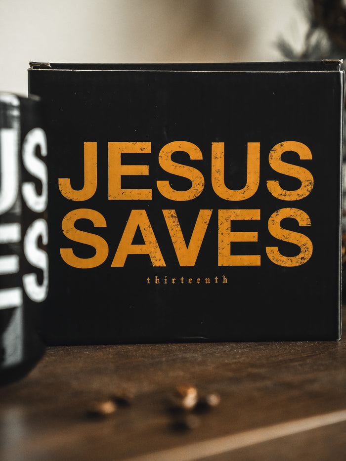 A black sign with large, bold yellow "JESUS SAVES" text, like the Jesus Saves Mug by 13th brand; "thirteenth" appears below in small letters. A similar-style object is partly seen at the left edge.