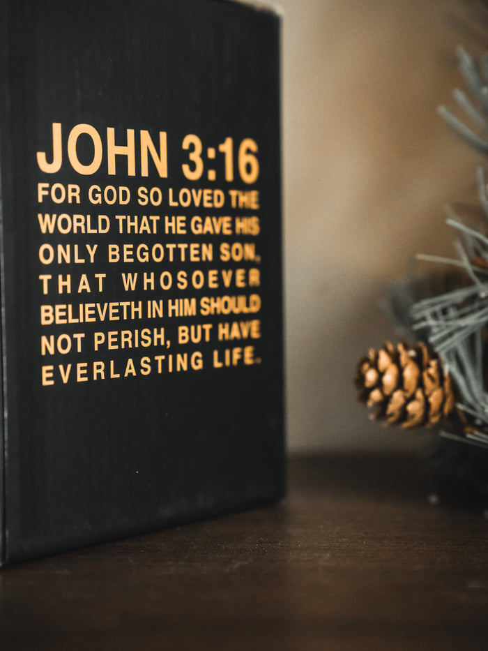 A close-up shows a black box with “John 3:16” in yellow beside a pinecone, an evergreen branch, and the 13th brand’s Jesus Saves Mug on a wooden surface.