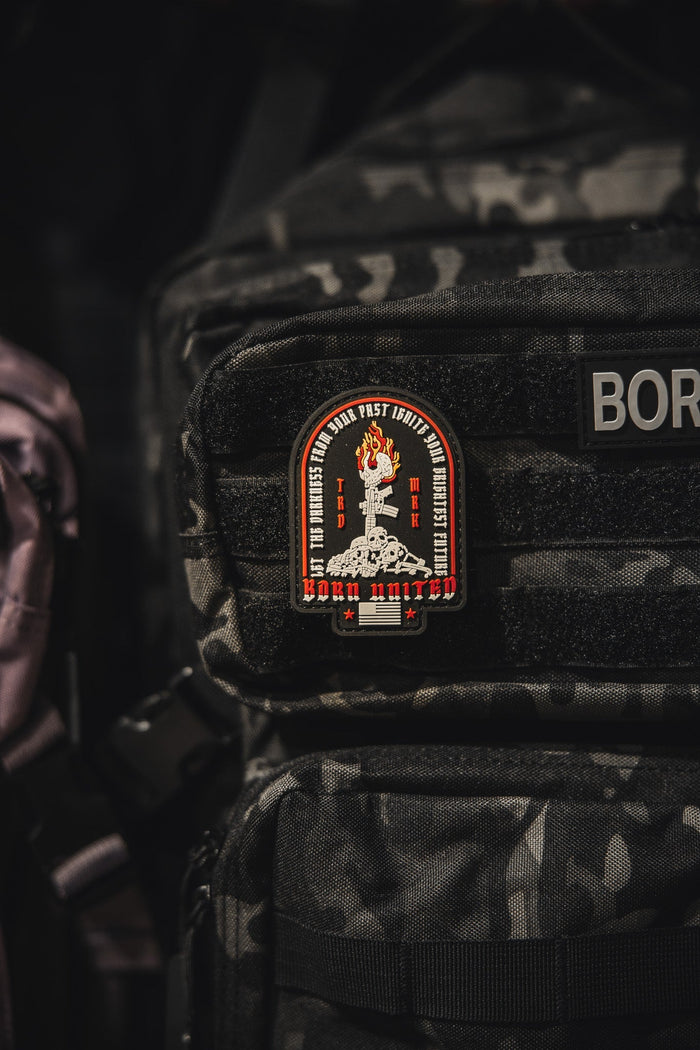 The Let The Darkness PVC Patch by Born United Private Label, featuring a flaming torch, red and white details, stars, and edge text, is attached to the front pocket of a black camo-patterned backpack.