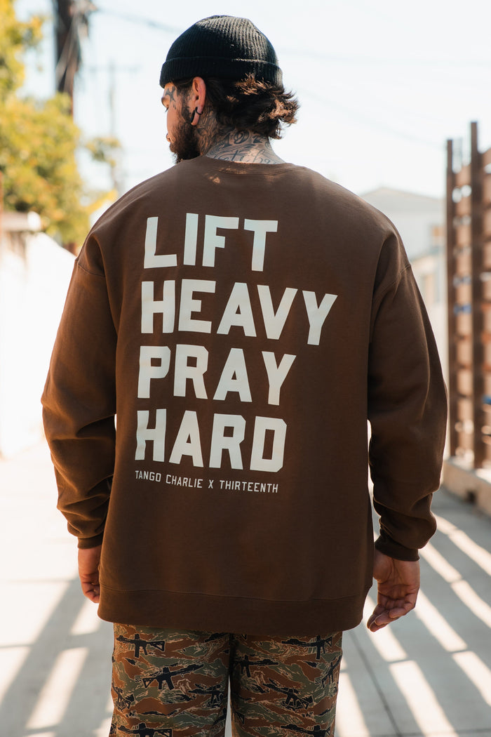 A man stands outdoors, facing away from the camera, wearing the Tango Charlie Apparel "Lift Heavy. Pray Hard." crewneck in brown with bold white text, paired with camo pants. Sunlight and a fence are visible in the background.