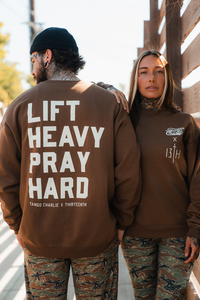 A man and woman stand outdoors in matching Tango Charlie Apparel camo pants and brown "Lift Heavy. Pray Hard. - Crewneck" sweatshirts, with his showing the bold slogan and hers featuring smaller white front logos.
