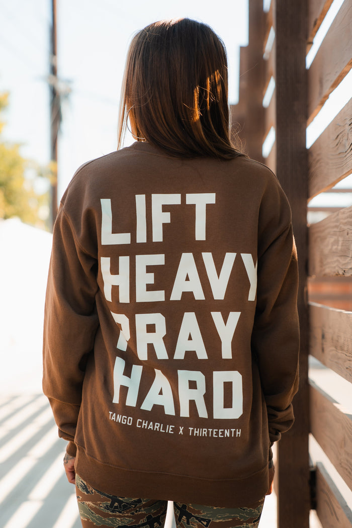 A person with long hair wears the Lift Heavy. Pray Hard. - Crewneck by Tango Charlie Apparel, featuring bold white text, while standing outdoors near a wooden fence—embodying Christian fitness.