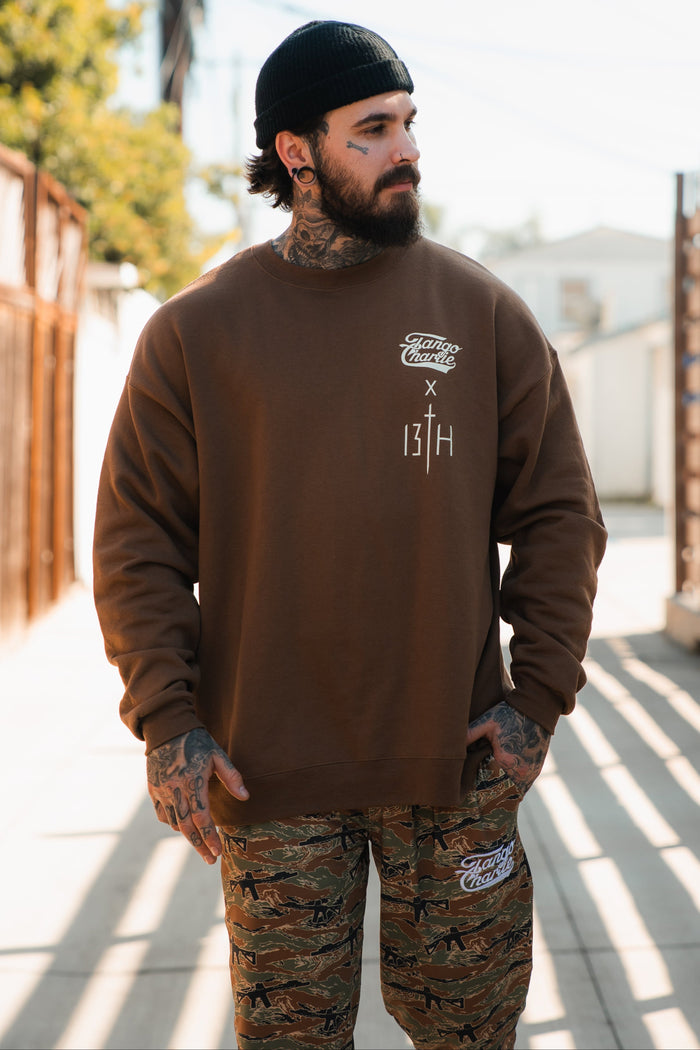 A tattooed bearded man in a black beanie and camo pants stands outdoors, wearing the "Lift Heavy. Pray Hard." crewneck by Tango Charlie Apparel, embodying resilience.