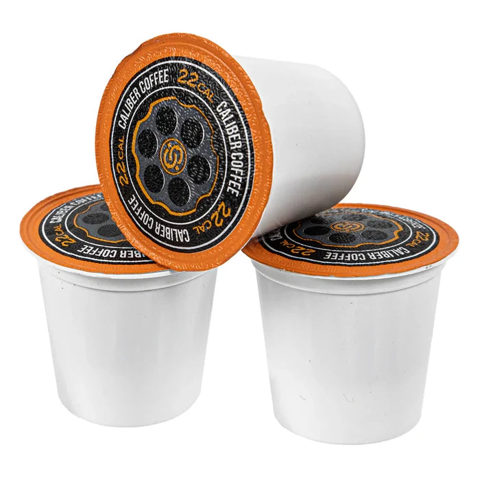 Three white single-serve pods with orange foil lids, labeled Caliber Coffee Company Light Roast .22 Cal Shells, are arranged neatly, with one leaning on the others. These pods are designed like a bullet chamber and provide a light roast specialty grade coffee experience.