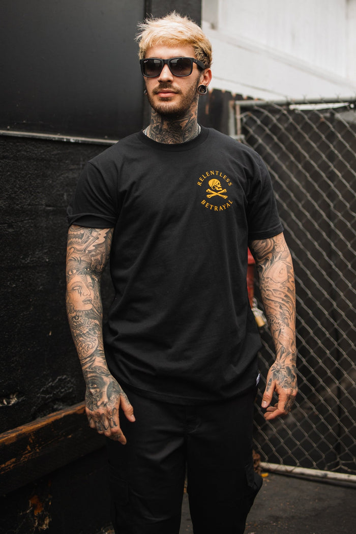 A tattooed man with sunglasses stands by a chain-link fence, wearing a black Lost At Sea T-shirt from Relentless Betrayal, featuring a skull and crossbones with Relentless Betrayal in bold yellow.