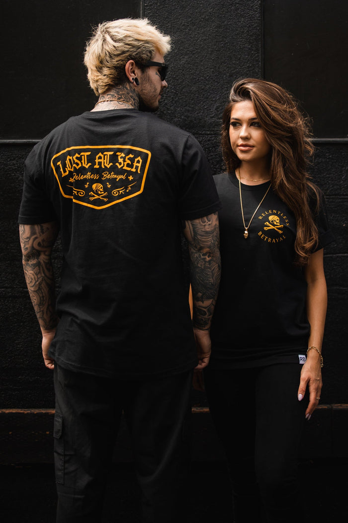 A man and woman stand against a dark background, both in Relentless Betrayals Lost At Sea black tees. The mans tattooed arm highlights the design on his shirts back, while the long-haired woman showcases it on her front. Crafted from fine jersey cotton, these unisex tees are a statement of style.