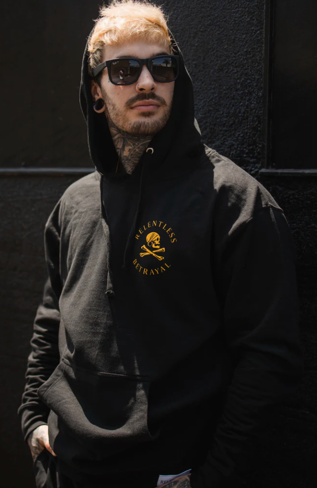 A man with sunglasses and tattoos wears a Lost at Sea Hoodie by Relentless Betrayal, made from a comfortable cotton/polyester blend featuring yellow text. He stands against a dark background with an edgy snapback complementing his style.