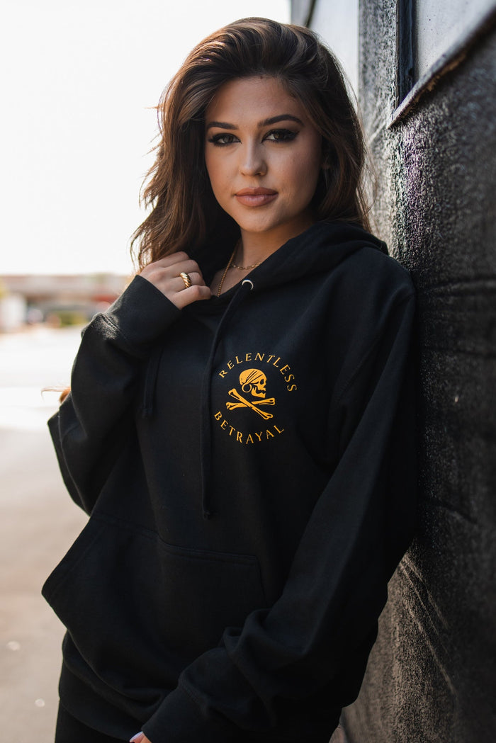 A woman with long hair is outdoors wearing a black Lost at Sea Hoodie by Relentless Betrayal. She leans against a wall, adjusting her unisex snapback while looking at the camera, sunlight emphasizing the soft cotton/polyester fabric.