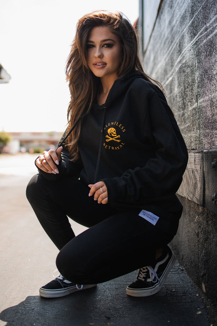Someone with long hair squats outdoors near a wall, wearing Relentless Betrayals Lost at Sea hoodie in black with yellow text, black pants, and black-and-white sneakers. They sport a snapback and look at the camera as sunlight highlights their face and part of the scene.