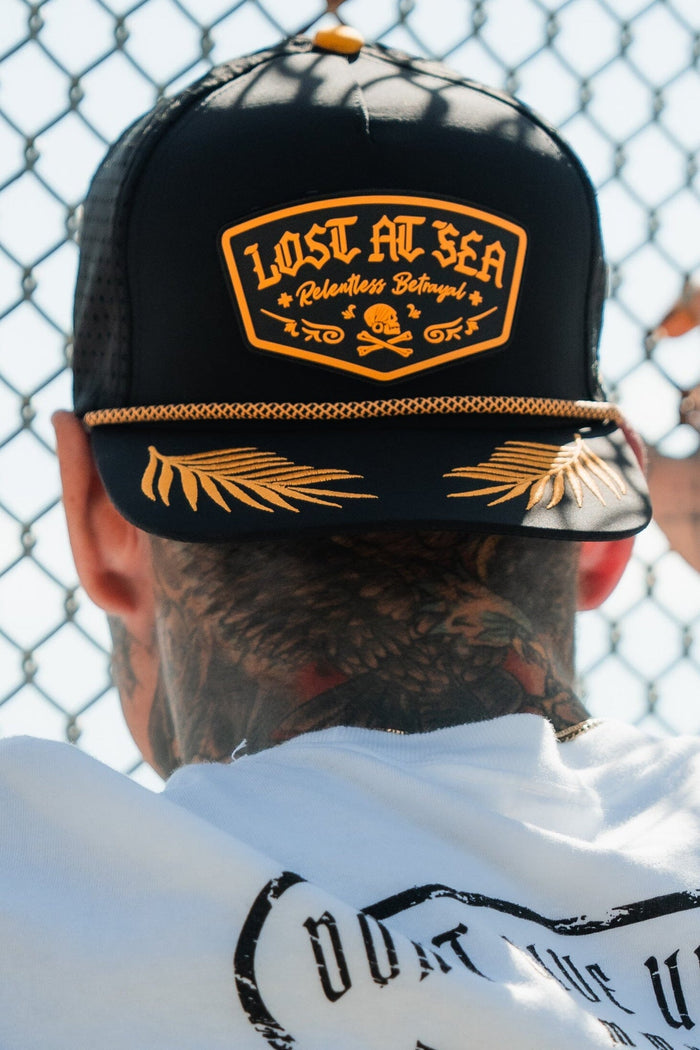 The image shows the back of a persons head wearing the Lost At Sea PVC Snapback by Relentless Betrayal, featuring yellow details and the text Lost at Sea Relentless Betrayal, set against a chain-link fence background.