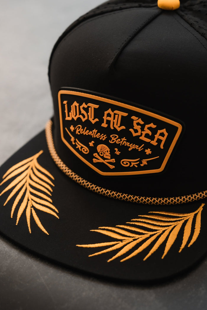 The Lost At Sea PVC Snapback by Relentless Betrayal is a black cap ideal for the pirate lifestyle, featuring a gold-embroidered design, a PVC patch with Lost at Sea and Relentless Betrayal, plus gold palm fronds and a braided chain on the brim.