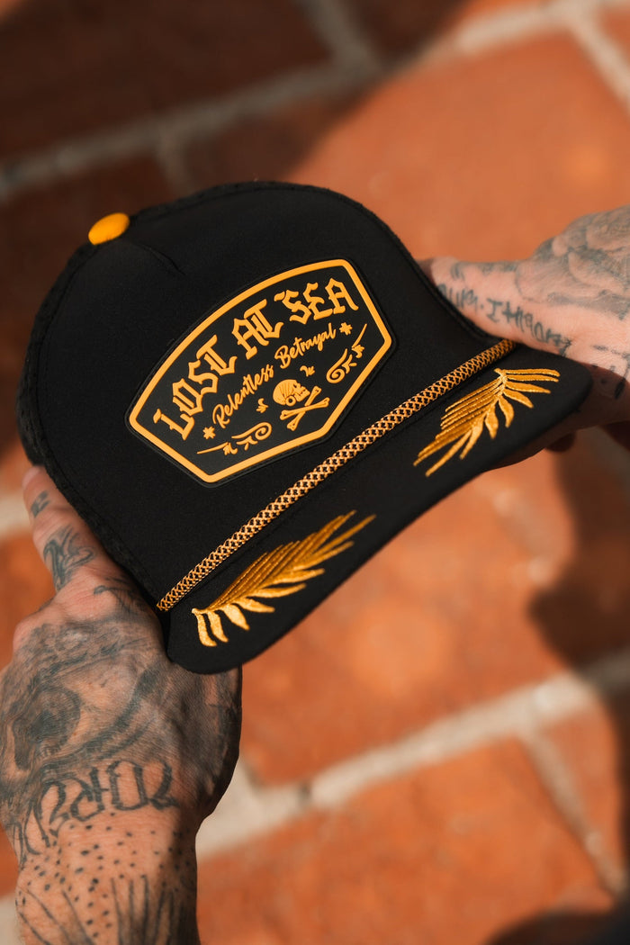 A tattooed hand holds the Lost At Sea PVC Snapback by Relentless Betrayal. This water-resistant black cap features a LOST AT SEA PVC rubber patch and yellow leaf embroidery on the brim, set against a tiled floor for a subtle pirate lifestyle vibe.