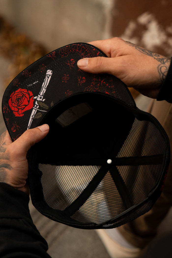 A person with tattooed hands holds the Relentless Betrayal Love/Hate 7 Panel Trucker Snapback, featuring a black mesh design. Its brim showcases a red rose and dagger on a dark background, while the interior highlights meticulous construction.