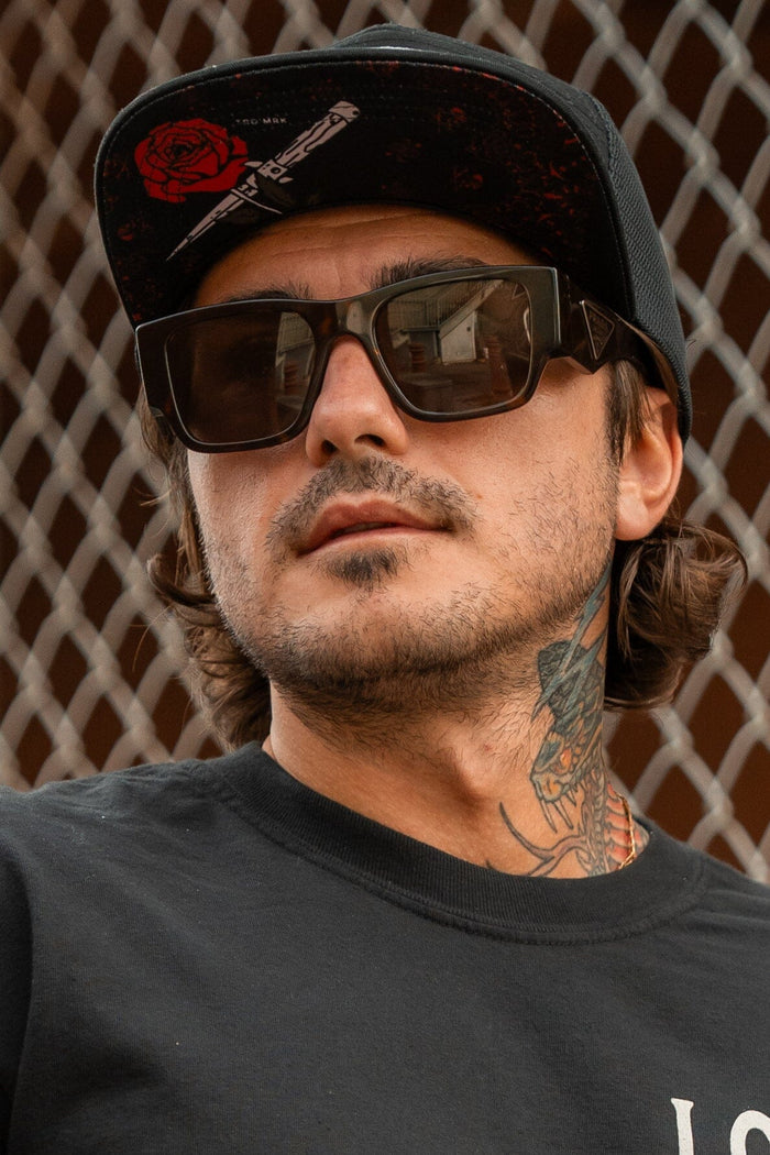 Someone in a Relentless Betrayal Love/Hate 7 Panel Trucker Snapback with a red design, large sunglasses, and a black T-shirt stands by a chain-link fence. They have neck tattoos and are gazing to the side.