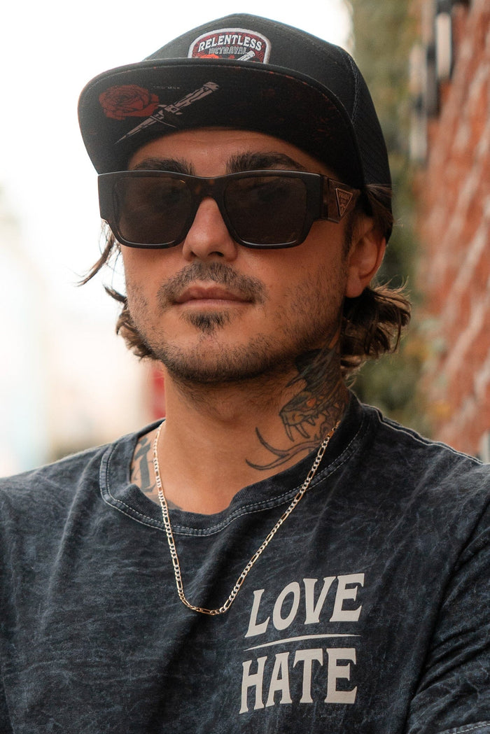 A man in a Relentless Betrayal Love/Hate 7 Panel Trucker Snapback, large sunglasses, and a silver necklace leans against a brick wall. His neck tattoos complement the shirt with bold LOVE and HATE lettering.