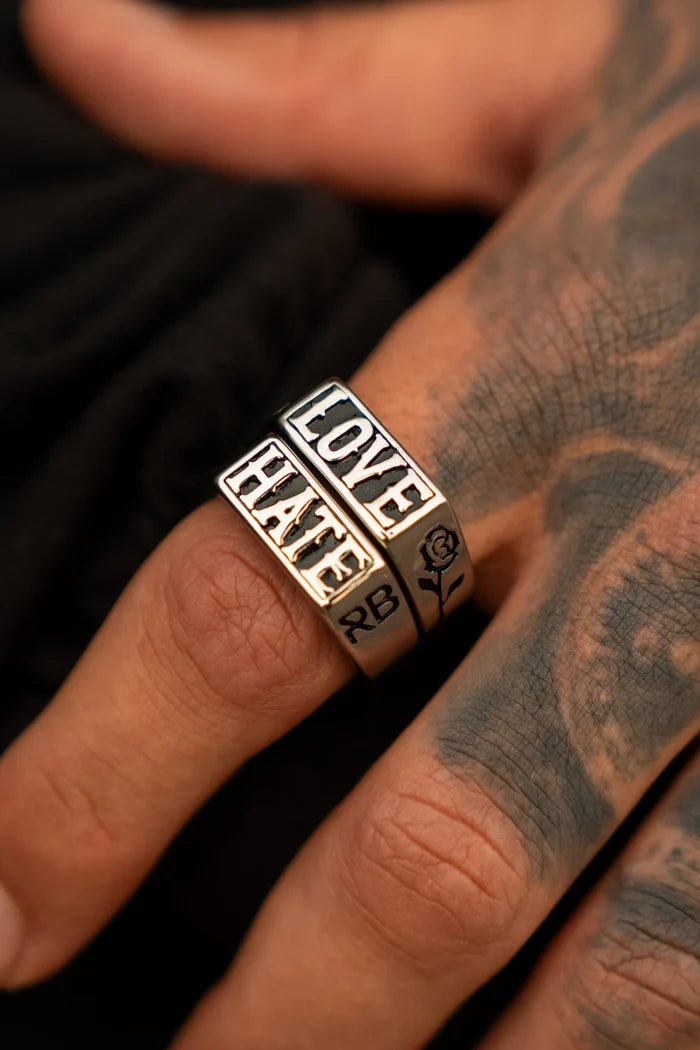 A hand showcases a duo from Relentless Betrayals Love/Hate Rings on the middle finger—one with LOVE and one with HATE, both in stainless steel. Detailed tattoos, like a rose, add artistry against a stark black backdrop.
