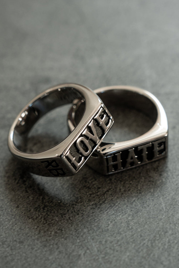 Two silver rings lie on a gray surface: one is engraved with LOVE and the other with HATE. This handcrafted designer duo, from Relentless Betrayals Love/Hate Rings collection, forms an overlapping design.