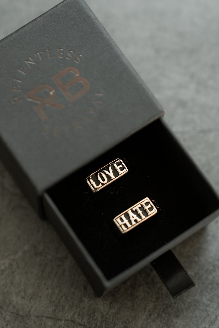 A pair of handcrafted silver rings, engraved with LOVE and HATE, is displayed in an open black jewelry box on a gray surface. The box lid features indistinct embossed text from the brand Relentless Betrayal.