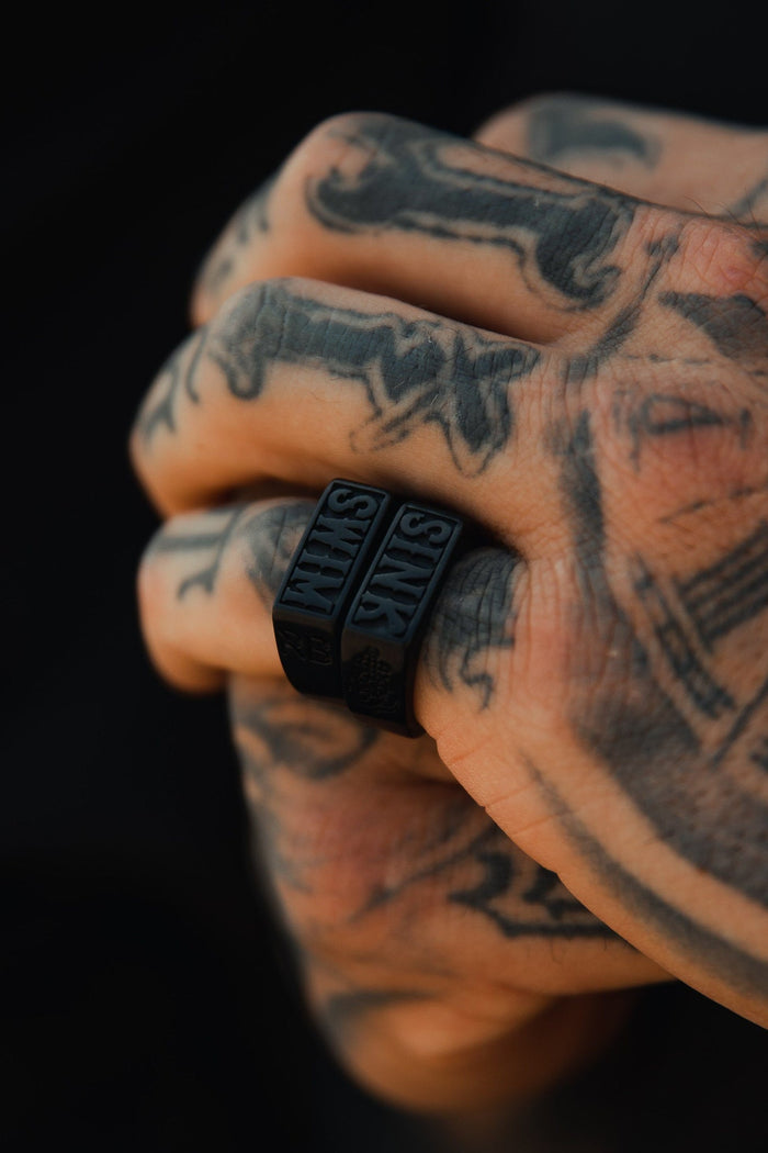 A tattooed hand forms a fist, showcasing two handcrafted Matte Black Sink or Swim Rings by Relentless Betrayal on the middle finger. The rings, featuring raised SINK letters, stand out against detailed tattoos in contrast to a blurred backdrop.