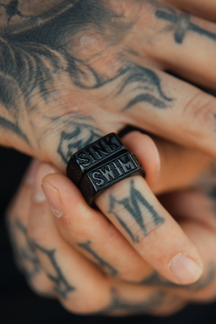 Close-up of tattooed hands, highlighting a finger wearing two Relentless Betrayals Matte Black Sink or Swim Rings with engraved text. The intricate tattoos provide an artistic touch to the image.