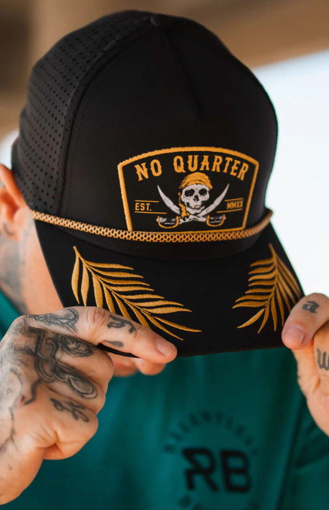 A person adjusts the Relentless Betrayal No Quarter Blackout Snapback, featuring a skull and crossbones with No Quarter and Est. MMIX embroidery and gold leaf patterns, atop their tattooed skin while wearing a green top.