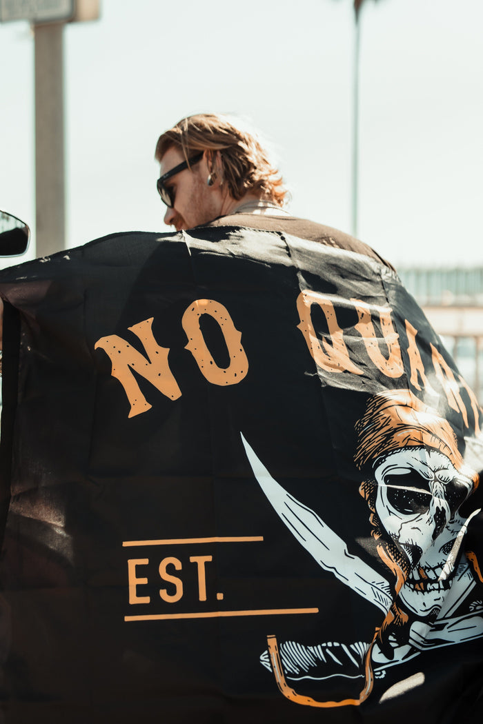 A person with long hair and sunglasses proudly holds the Relentless Betrayal No Quarter Flag, a durable polyester banner featuring a skull with a bandana, crossed swords, and bold NO QUARTER text, waving defiantly in the sunny outdoor scene.