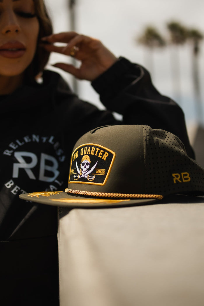 A person wears a Relentless Betrayal No Quarter Olive Snapback, water-resistant with yellow details and a skull and crossbones patch, against a blurred backdrop of palm trees and clouds.
