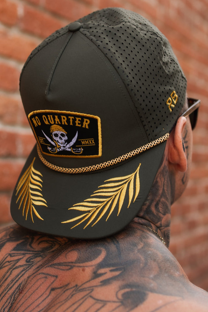 A tattooed person stands against a brick wall, wearing an olive No Quarter snapback by Relentless Betrayal, featuring water-resistant fabric, perforated mesh, a pirate-themed patch, and embroidered gold leaves on the back.