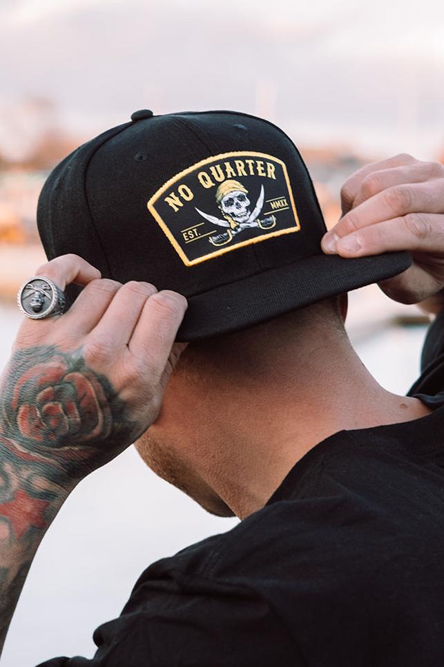 A person, adorned with tattoos and rings, adjusts a No Quarter Snapback from Relentless Betrayal featuring a skull and crossbones logo. Their focus on the flat-brim hat stands out against the blurred background.