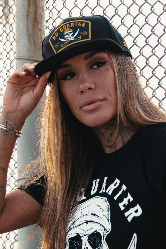 A person with long brown hair and tattoos adjusts a Relentless Betrayal No Quarter Snapback adorned with a skull emblem. They wear a matching T-shirt while standing in front of a chain-link fence.