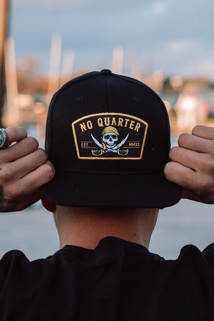 A person holds the brim of a No Quarter Snapback by Relentless Betrayal, featuring a yellow and black patch with crossed swords and a skull reading No Quarter EST. MMXX, against a blurred background.