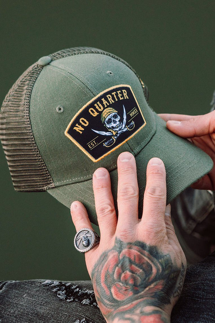 A tattooed hand, with a rose design, holds the green No Quarter Trucker Snapback from Relentless Betrayal. The cap features a skull and crossed swords patch with NO QUARTER and EST. MMXII. A person adjusts the snapback while wearing a silver ring.