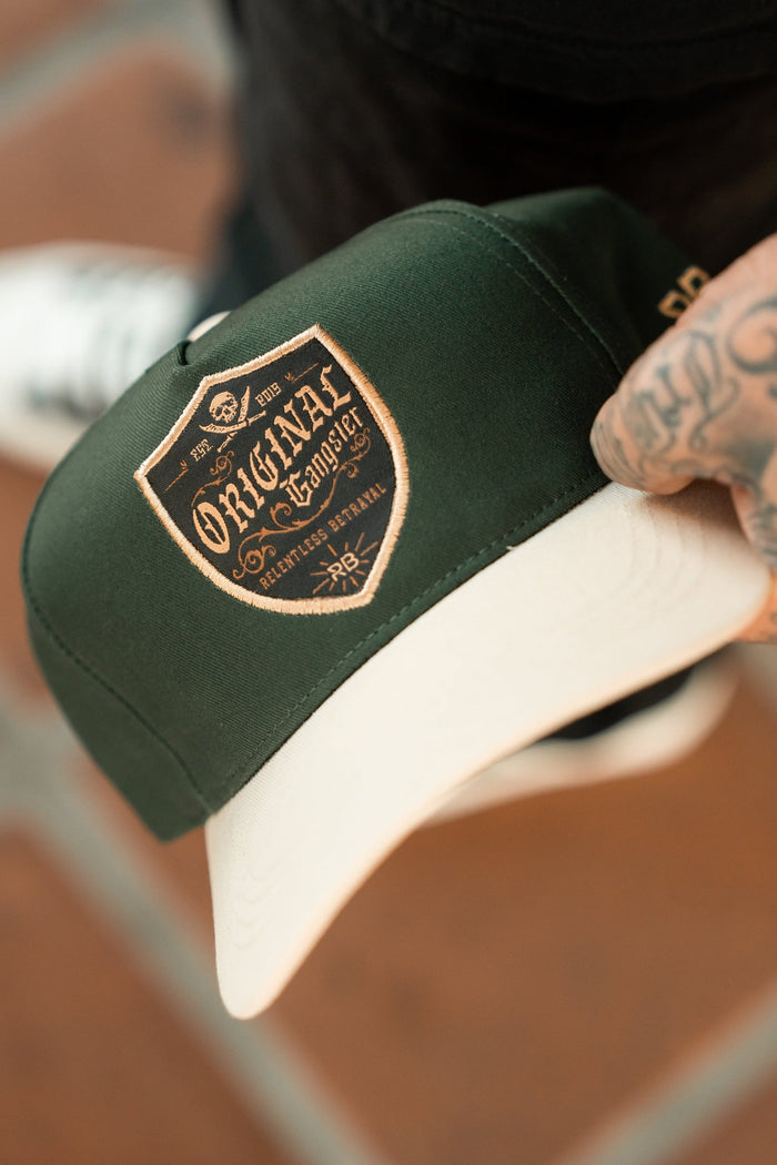 A tattooed person holds the Relentless Betrayals OG Forest Green Snapback hat, featuring a beige brim and a shield-shaped Original logo with intricate designs. Theyre wearing dark pants and white shoes.