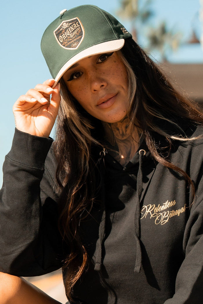 A person with long hair and tattoos wears a black hoodie emblazoned with Relentless Betrayal and an OG Forest Green Snapback. They adjust their cap outdoors against a backdrop of blurred trees and sky.