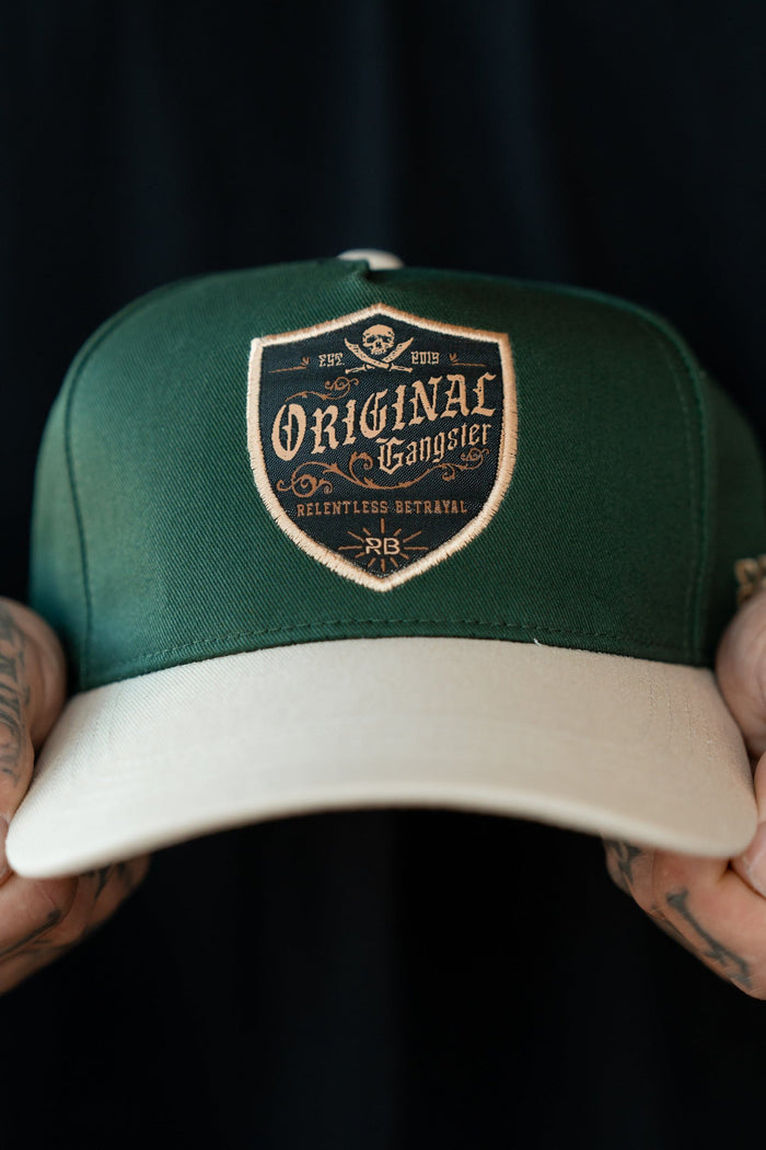 Close-up of tattooed hands holding an OG Forest Green Snapback by Relentless Betrayal. The cap, featuring a beige brim, showcases a patch with Original Gangster in ornate lettering and a small emblem above, set against a dark background.