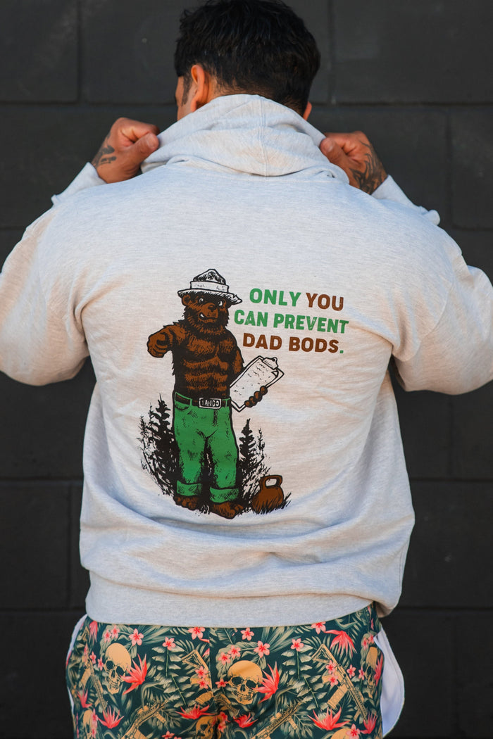 A person faces away from the camera, lifting the hood of Tango Charlie Apparel’s Only You Can Prevent Dad Bod Hoodie—gray with a bear illustration and humorous text—and wears colorful skull-patterned shorts.